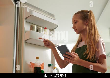 Preteen girl with shopping list on smartphone checking shelves in fridge Stock Photo