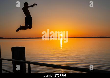 Boy jumps from the wooden jetty into the water at sunset, Chieming, Chiemsee, Chiemgau, Bavaria, Germany Stock Photo