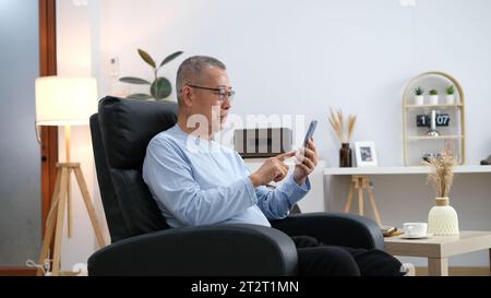 Happiness of wellness elderly asian man with white hairs sitting on sofa using mobile phone at home. Stock Photo