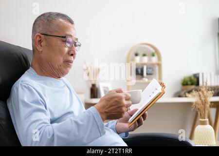 elderly man in eyeglasses sitting at home on the sofa reading a book and holding a cup of tea or coffee in his hand. Stock Photo