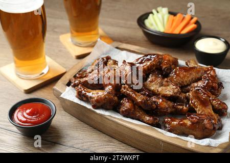 Tasty chicken wings, sauces, vegetable sticks and glasses of beer on wooden table, closeup. Delicious snacks Stock Photo