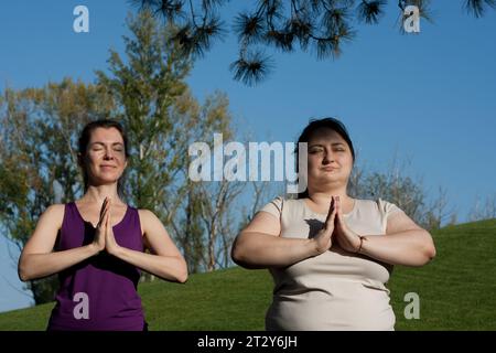 Two middle-aged women practice yoga in city park, meditate, standing, sun salutation, namaste. Healthy lifestyle, fitness, yoga, pilates outdoors. ove Stock Photo