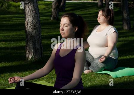 Two middle-aged women practice yoga in city park, meditate sitting on mats on grass in sun. Healthy lifestyle, fitness, yoga, pilates outdoors. overwe Stock Photo