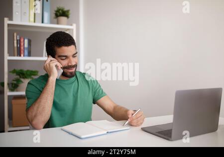 Cheerful millennial arab man manager at table with laptop working remotely, call by phone Stock Photo