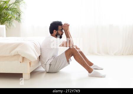 Life Troubles Concept. Depressed Young Indian Man Sitting On Floor Near Bed Stock Photo