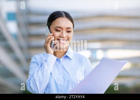 Cheerful Asian Businesswoman Talking On Cellphone Holding Papers Outside Stock Photo