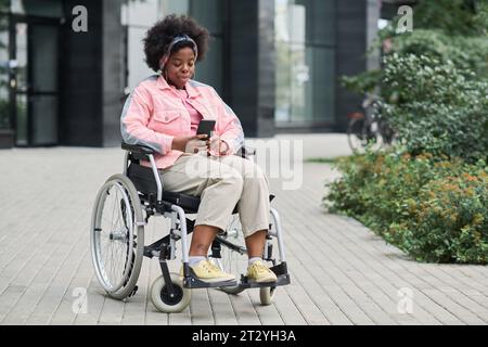 African American woman with disability sitting on wheelchair and using smartphone outdoors Stock Photo