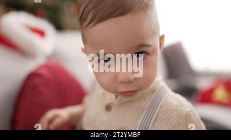 Adorable toddler standing by christmas tree with serious expression at home Stock Photo