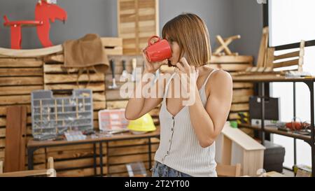 Serious young hispanic female carpenter sipping morning coffee, engaging in professional business call at bustling carpentry workshop Stock Photo