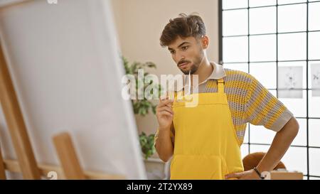 Serious young arab man, a talented artist, holding glasses, deep in thoughts, looking at his masterpiece, drawing attentively at art studio, embracing Stock Photo