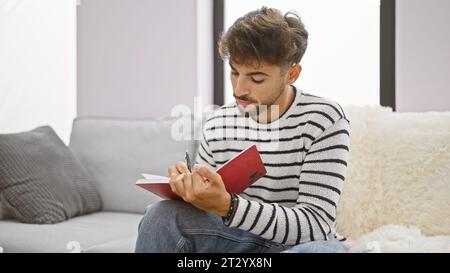 Smart young arab man, comfortably seated on sofa, intensely studying literature, focused on writing notes indoor at his home living room, a serene ind Stock Photo