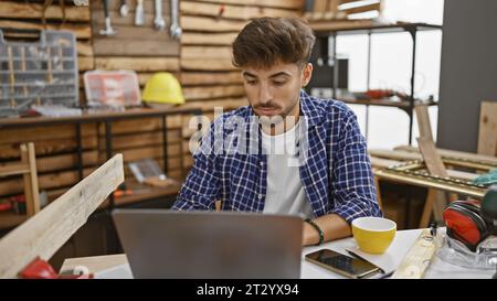 Handsome young arab man, professional carpenter, using laptop at his timber workshop. engaged in his woodworking business, masterly shaping furniture. Stock Photo