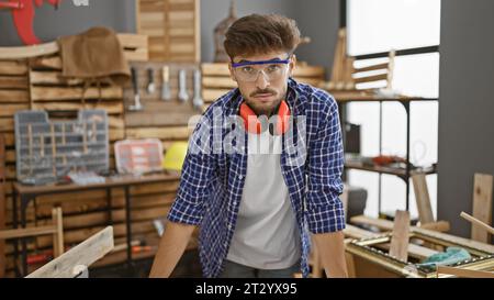 In the heart of the carpentry industry, a serious-faced young man, an arab carpenter wearing glasses, breathes in the essence of woodwork amidst the b Stock Photo