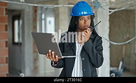 African american woman architect using laptop talking on walkie talkie at construction site Stock Photo
