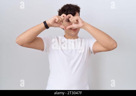 Young non binary man wearing casual white t shirt doing heart shape with hand and fingers smiling looking through sign Stock Photo
