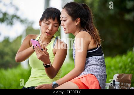 two young asian women female friends in sportswear relaxing chatting sharing cellphone photos outdoors Stock Photo