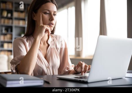 Pensive woman sitting at desk using laptop surfing internet Stock Photo