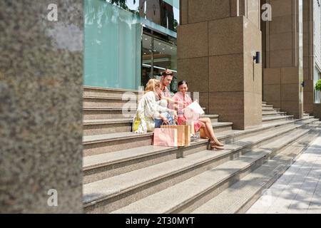 Cheerful friends sitting on steps of shopping mall and discussing purchases they made on sale Stock Photo