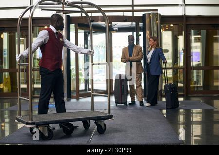 Doorman suggesting the cart for guests while they standing with luggage near the entrance Stock Photo