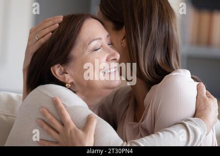Adult daughter embraces aged 50s mother seated on sofa indoors Stock Photo