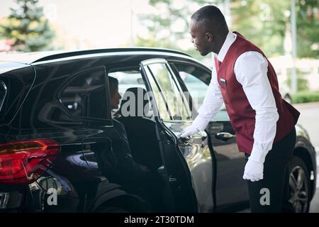 African American doorman opening the door of the car for guest and meeting her outside Stock Photo