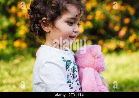 A curly-haired young girl clutches a pink plush toy, lost in thought amidst an autumnal park setting. The warm sunlight enhances the nostalgic and inn Stock Photo