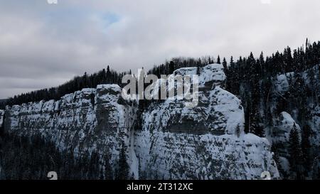 Aerial winter snow scene with ice covered cliff. Clip. White cliff edge with forested top. Stock Photo