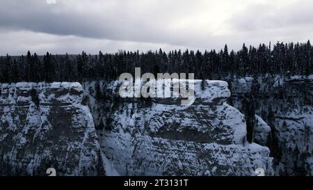 Aerial winter snow scene with ice covered cliff. Clip. White cliff edge with forested top. Stock Photo