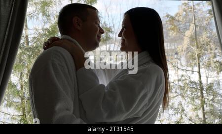 Loving man and woman are hugging in morning in bedroom against window. Action. Husband and wife near window with trees behind. Stock Photo