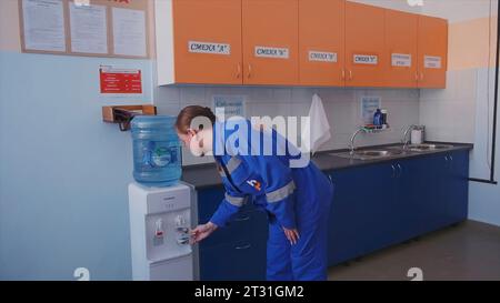 Serbia, Belgrade - June 20, 2023: Woman in overalls at factory. Clip. Woman in work suit goes to drink water. Kitchen area at factory for workers Stock Photo