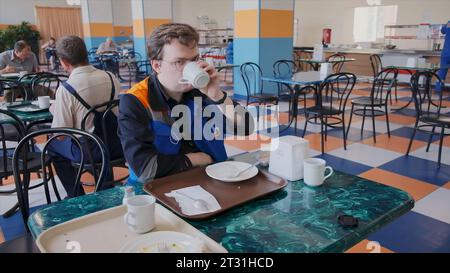 Serbia, Belgrade - June 20, 2023: Worker in uniform eats in dining room. Clip. Young man in cafeteria drinks coffee on work shift. Cafeteria at Stock Photo