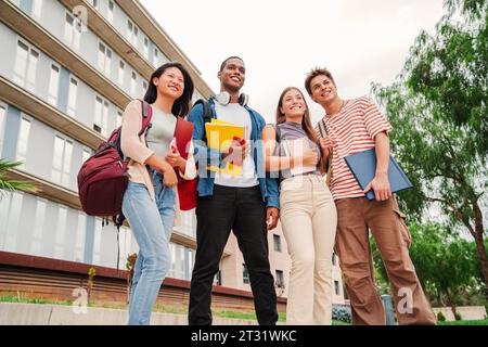 Portrait of a group of smiling multiracial university students carrying backpacks and folders, standing at campus. Multiracial young classmates socializing after class. Teenage guys and young ladies. High quality photo Stock Photo