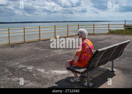 An elderly woman sitting on a bench looking out to sea Stock Photo