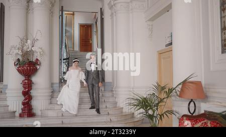 Man and bride go down stairs. Action. Newlyweds in outfits descend from registry office. Bride and groom go down stairs of registry office Stock Photo