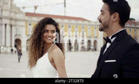 Young and beautiful couple walking along historic buildings and talking. Action. Man in suit and woman in white dress holding hands in the city square Stock Photo