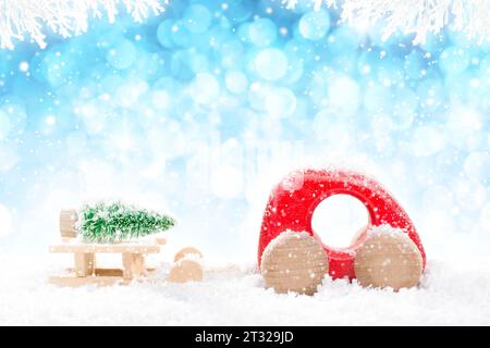 Red Wooden Car Pulling Sled with Green Christmas Tree in perspective on bright blue bokeh winter background with white snowy branches and falling snow Stock Photo