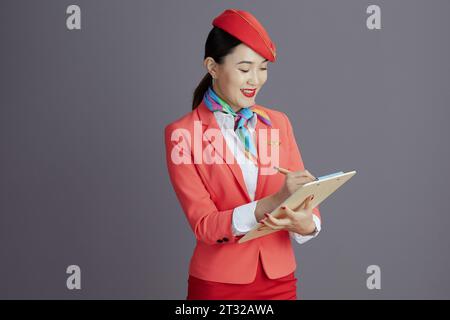 smiling stylish asian female flight attendant in red skirt, jacket and hat uniform with clipboard isolated on grey. Stock Photo