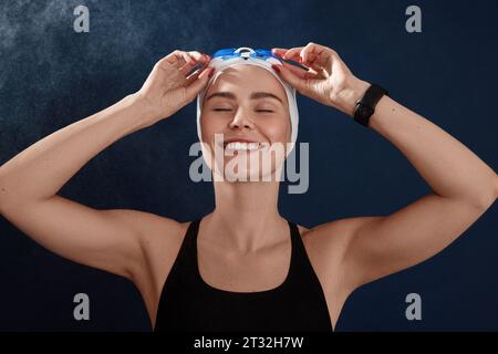 Young smiling female swimmer with closed eyes adjusting goggles standing over a blue backdrop Stock Photo