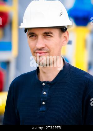 Portrait of Caucasian builder or gas industry engineer wearing white construction helmet. Confident looking working man aged 35-40 stands against background of yellow pipes. Reliable contractor. Stock Photo