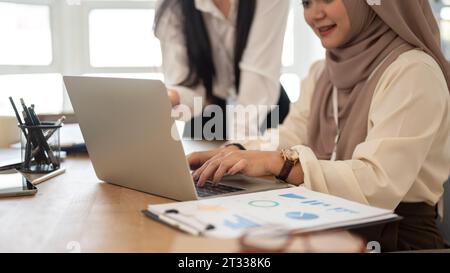 Close-up side view image of a professional Asian-Muslim businesswoman using her laptop while discussing work with her colleague in the meeting room. Stock Photo