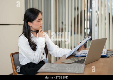 A concentrated and thoughtful Asian businesswoman or female office employee is examining business financial report, working at her desk in a modern of Stock Photo