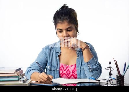 A pretty young Indian college student studying and biting finger on study table and white background Stock Photo