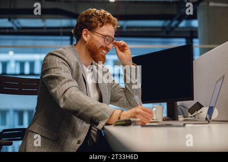 Smiling businessman working on laptop and making notes while sitting desk on office background Stock Photo