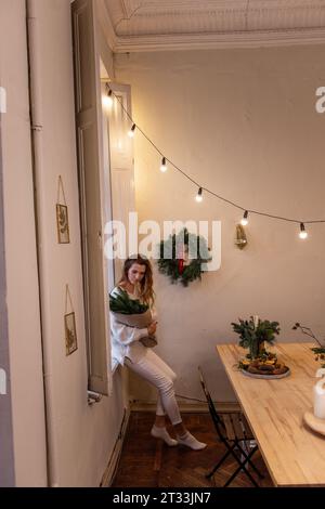 Vertical shot of stylish young woman in white knitted sweater, sitting on windowsill, holding in hands eco-friendly winter bouquet, spruce branches in Stock Photo