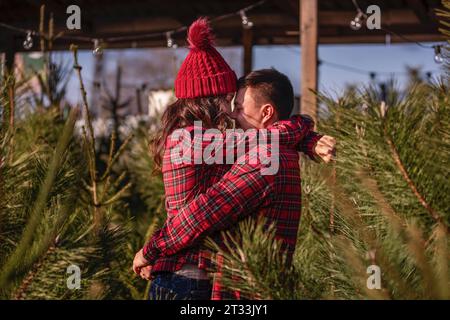 Beautiful couple in love in checkered red shirts, knitted hats hug tightly nose to nose face to face among green Christmas tree market. Young man laug Stock Photo