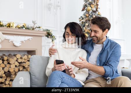 Happy young couple man and woman sitting on sofa together, hugging and watching video online using app on phone, joyful choosing shopping in online store, celebrating christmas and new year. Stock Photo