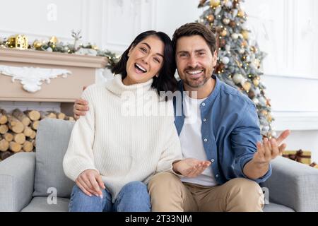 Webcam view, online video ringing, young couple man and woman looking at camera, talking with friends, congratulating on Christmas and New Year, sitting on winter day in living room, near gifts. Stock Photo