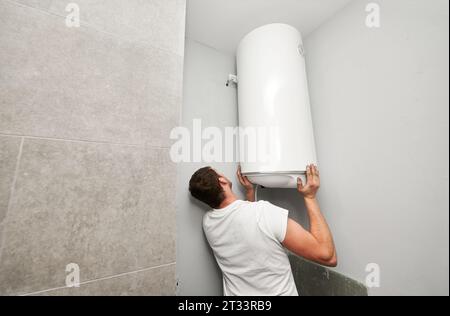 Back view of male worker installing boiler heating system in bathroom at home. Man plumber hanging gas boiler on the wall in apartment under renovation. Plumbing works and home refurbishment concept. Stock Photo