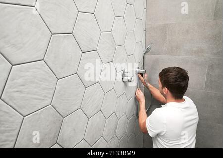 Man plumber installing handheld showerhead in bathroom. Male worker fixing metal bath showerhead and doing plumbing works while working on home renovation. Stock Photo