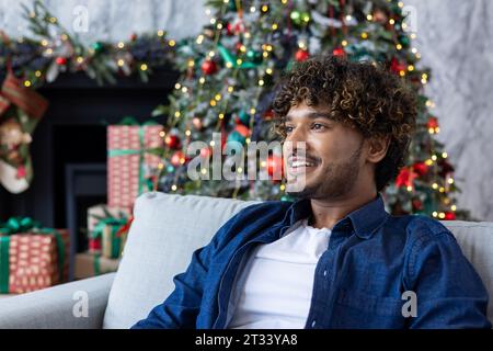 Close-up portrait of a young man for Christmas, hispanic man smiling and looking away from the window , sitting on the sofa in the living room near the Christmas tree, celebrating the new year. Stock Photo